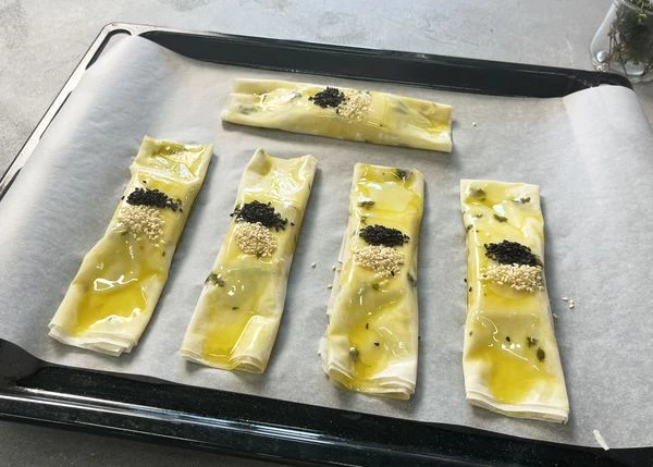 Переложить на противень, застеленный пергаментом. Каждый конвертик смазать маслом, посыпать кунжутом. Выпекать в разогретой до 200 градусов духовке в течение 15-20 минут.