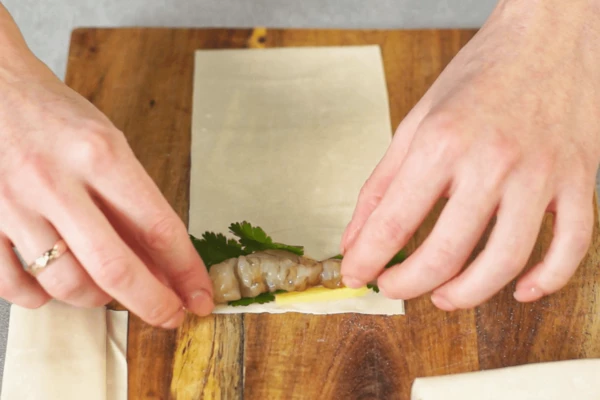 Имбирь очистить, нарезать длинной соломкой.
Тесто фило (2,5 листа) разморозить на нижней полке холодильника, нарезать на полоски.
Кинзу вымыть, обсушить, удалить стебли.
Креветки промакнуть от излишков соуса.
Полоску теста фило выложить на доску. На тесто выложить креветку, нарезанный имбирь и веточки кинзы.
