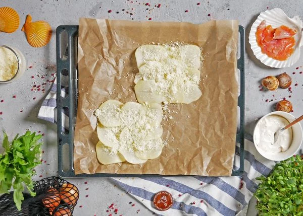 Картофель очистить, нарезать тонкими слайсами, промыть в холодной воде. На противень застелить пергамент, выложить картофель, сбрызнуть маслом, посолить. Обильно посыпать пармезаном.