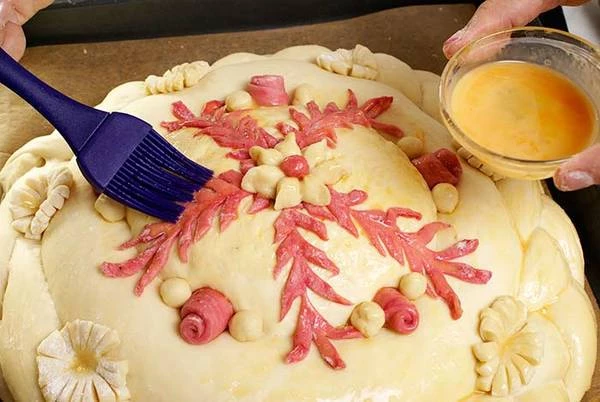 Вырезать различные украшения из тонко раскатанного теста : косички, веточки, цветочки и "приклеить" с помощью яичного белка к караваю. Потом смазать всю поверхность  взбитым яйцом.