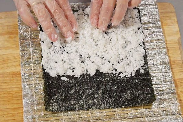 Водоросли нори положить вертикально, шершавой стороной вверх. Распределить равномерно рис, оставив сверху полоску ~2 см.