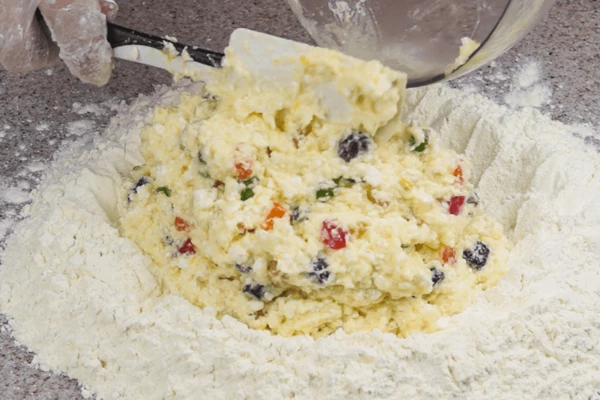 На стол горкой высыпать муку (можно делать замес в глубокой миске), добавить разрыхлитель, перемешать. Затем в центр выложить масляно-творожную массу и замесить пластичное тесто. Тесто должно быть мягким, но не расплываться.