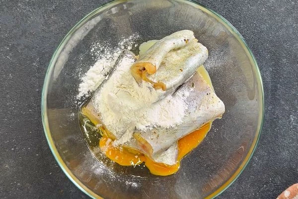Маринованный минтай переложить в другую емкость, добавить муку и яйцо. Запанировать кусочки рыбы со всех сторон.