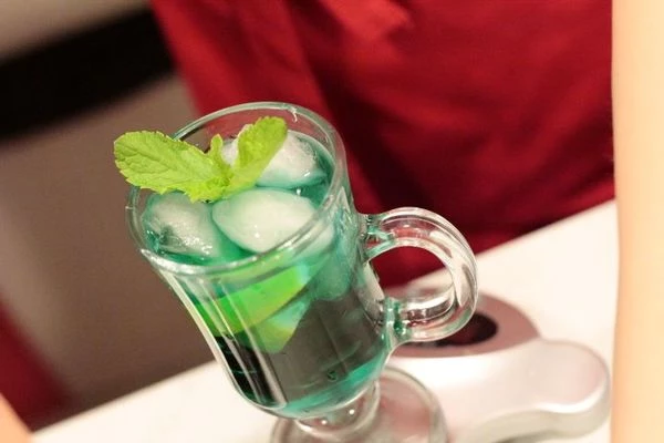 В бокал для коктейля влить сироп Блю Курасао и сироп Мята, добавить 1-2 дольки лайма.