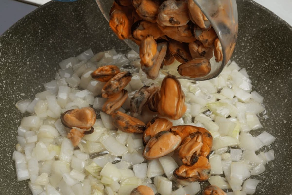 На разогретой с растительным маслом сковороде обжарить лук и чеснок, добавить размороженные мидии. Прогреть 2-3 минуты.