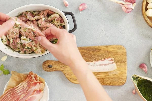 На доску выложить ломтик бекона на него выложить замаринованную свинину, свернуть в рулетик. 