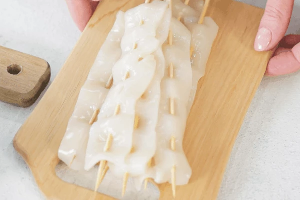 Кальмар разморозить на нижней полке холодильника, очистить, нарезать полосками, нанизать на деревянные шпажки.