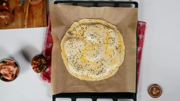 Тесто разморозить, раскатать, вырезать круг. Перенести его на томаты. Края теста прижать вилкой. Смазать взбитым яйцом. Посыпать кунжутом. Выпекать в разогретой до 190 градусов духовке 15 минут. Затем перевернуть и выпекать еще 7 минут. 