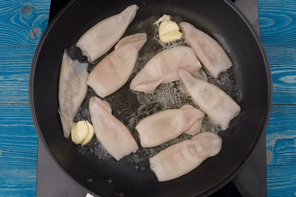 На сковороде разогреть смесь растительного и сливочного масла, добавить 2 зубчика раздавленного чеснока, обжарить тушки кальмара небольшими порциями до румяной корочки 1-2 минуты. Удалить чеснок, чтобы он не дал лишнюю горечь.