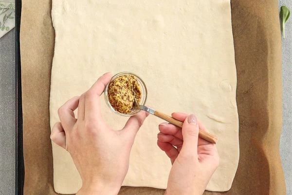 На него выложить размороженный пласт слоеного теста (слегка раскатанный). На тесто по центру намазать горчицу. 