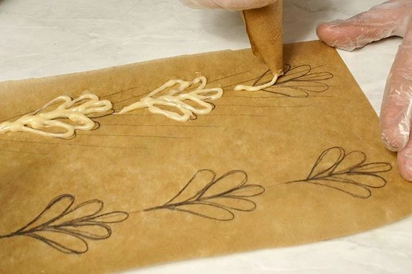 Переложить тесто в кондитерский мешок (или сделать кулек из пергамента). На листе пергамента нарисовать трафарет  карандашом, затем по готовому рисунку (с обратной стороны бумаги, чтобы карандаш не отпечатался на изделии) нарисовать узоры тестом. Выпечь в разогретой до 200С духовке до румяного цвета.