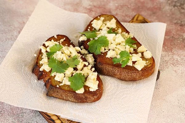 Обжаренный хлеб выложить на салфетку. Каждый ломтик натереть зубчиком чеснока.
Брынзу измельчить в крошку, выложить на обжаренные тосты, оформить листочками кинзы.
