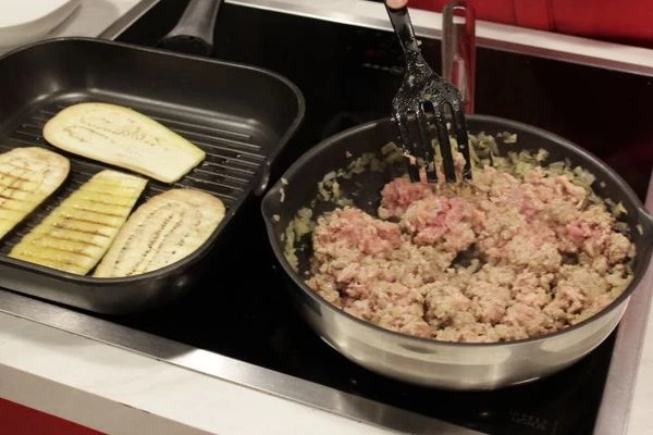 На разогретой с оливковым маслом сковороде обжарить лук и чеснок до прозрачности, добавить фарш, жарить еще 10 минут.  Одновременно на другой сковороде обжарить баклажаны с двух сторон до золотистого цвета, переложить на бумажное полотенце.