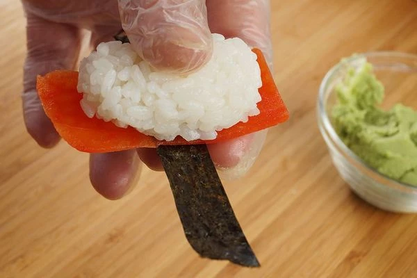 На ладонь положить полоску нори, затем ломтик сладкого перца, на перец колобок риса.
