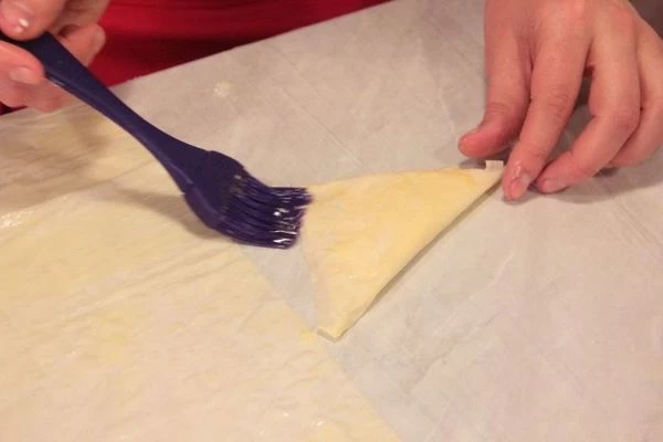 На каждую полоску с краю выложить начинку, завернуть в форме треугольника. Еще раз смазать маслом.