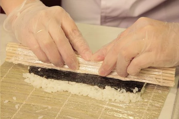 Коврик Макису оборачиваем пищевой пленкой. Выкладываем половинку нори горизонтальной полоской к себе шершавой стороной вверх.