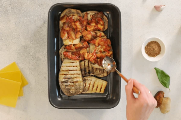 В форму для выпечки выложить слайсы баклажанов, распределить по ним с грибы и томатами.