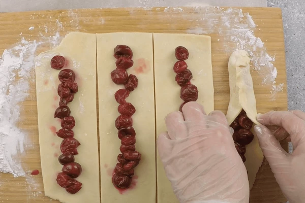  На доску насыпать крахмал, раскатать тесто в тонкий (2-3 мм) прямоугольный пласт, разрезать на 4-5 широких полоски. На каждую выложить вишню.