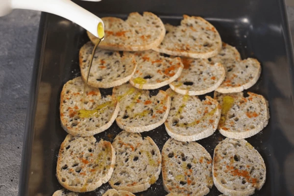 Багет нарезать тонкими слайсами, выложить на противень, посыпать паприкой, солью, полить оливковым маслом. Запекать при 160 градусах 3 минуты, перевернуть слайсы, запекать еще 3 минуты.
